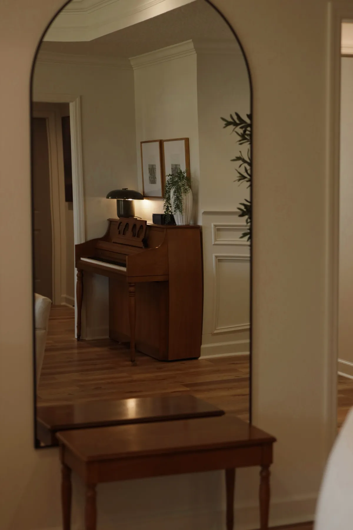 Tall arched mirror reflecting a hallway with an upright piano and wood console