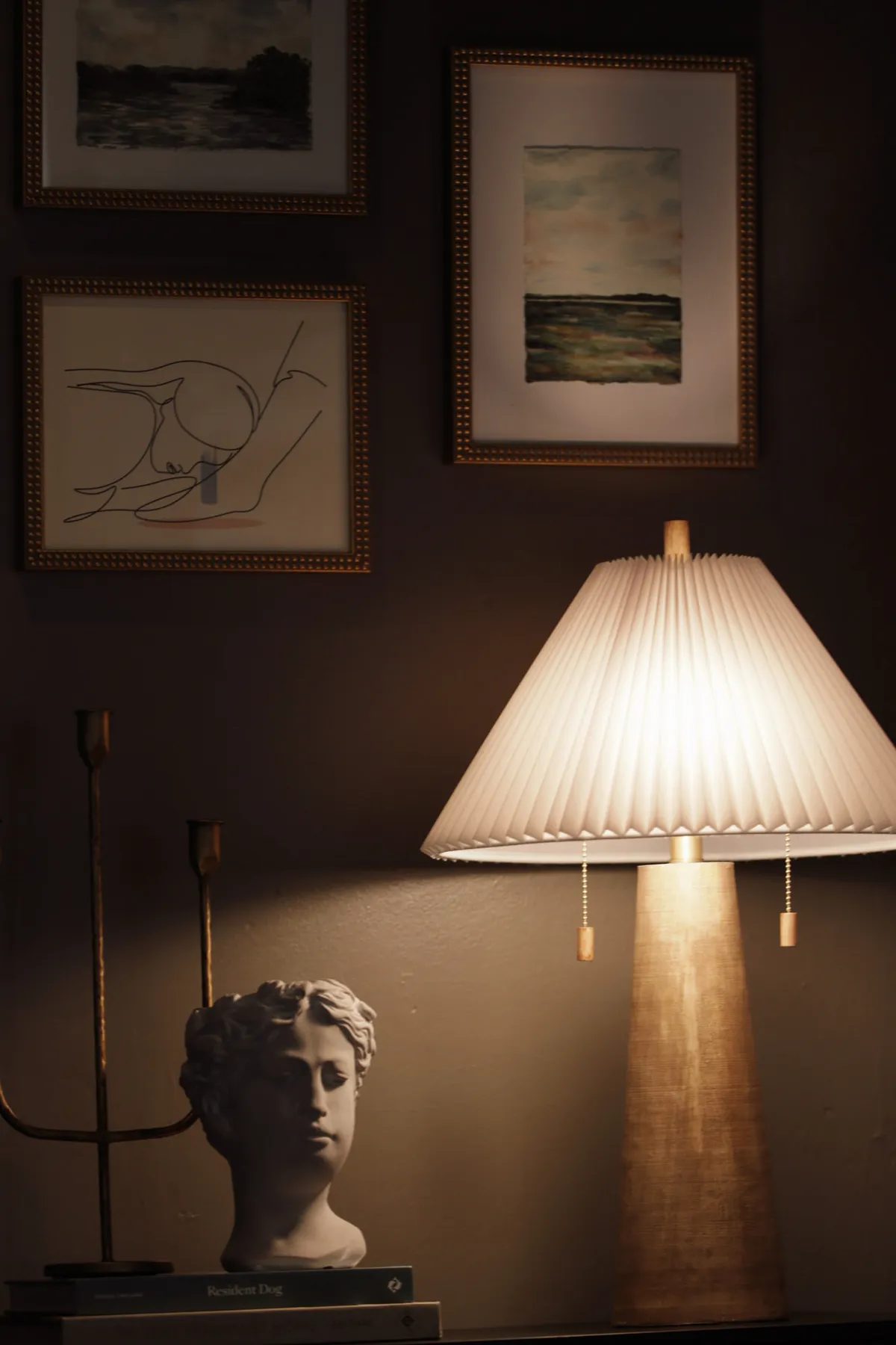 Warm table lamp beside a classical bust against a dark wall of framed art