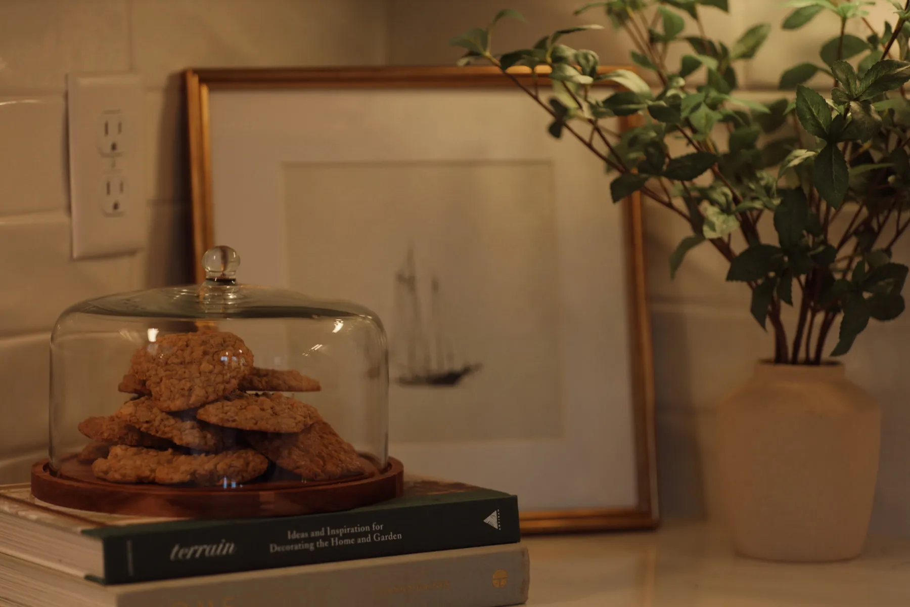 Glass cloche of cookies on a stack of books beside a framed sailboat print